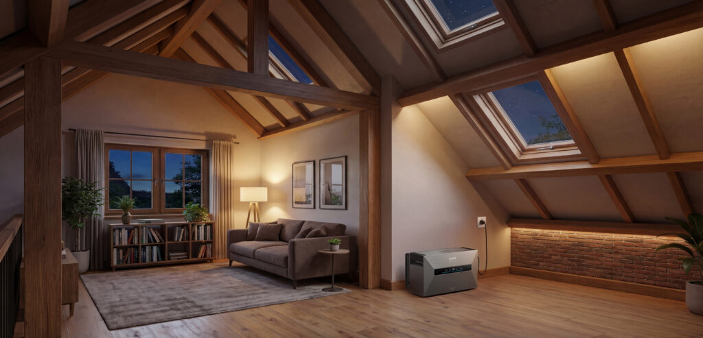 Tragbare Anker-Powerstation, die als kleine Heimbatterie dient, mit grauem Gehäuse in einem stimmungsvollen Dachzimmer mit Holzbalken, Dachfenstern und warmer Beleuchtung neben einer Backsteinwand, angeschlossen an die Steckdose für eine netzunabhängige Stromversorgung.